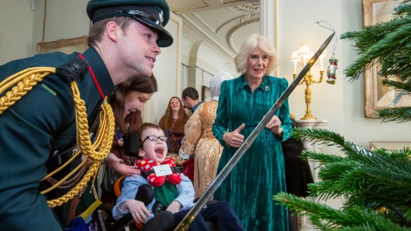 A man in a green and gold uniform using a sword to put a decoration on a Christmas tree. He is watched by a smiling young boy and the Queen, who is wearing a green dress.