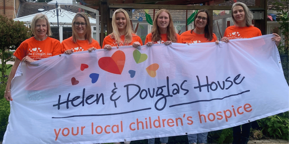 A photo of six women smiling as they hold up a Helen & Douglas House banner.